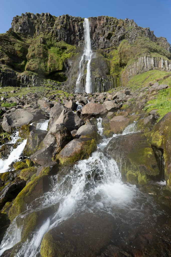 Bjarnarfoss Waterfall on a sunny day, with some rocks in the foreground.