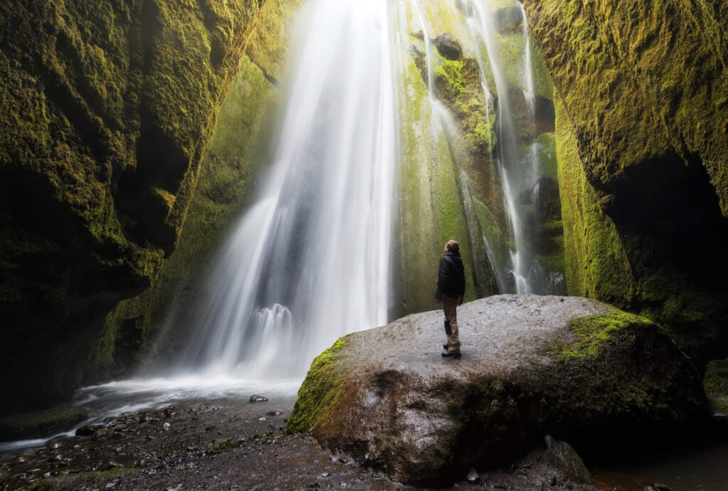 Person standing in a cave by a waterfall called Gljufrabui (or Gljúfurárfoss), looking at it