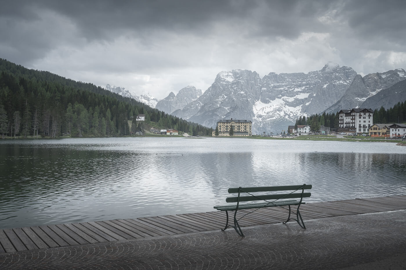 View of a bench by a lake in the Dolomites, with snowy mountains in the background, on a dark and gloomy day.