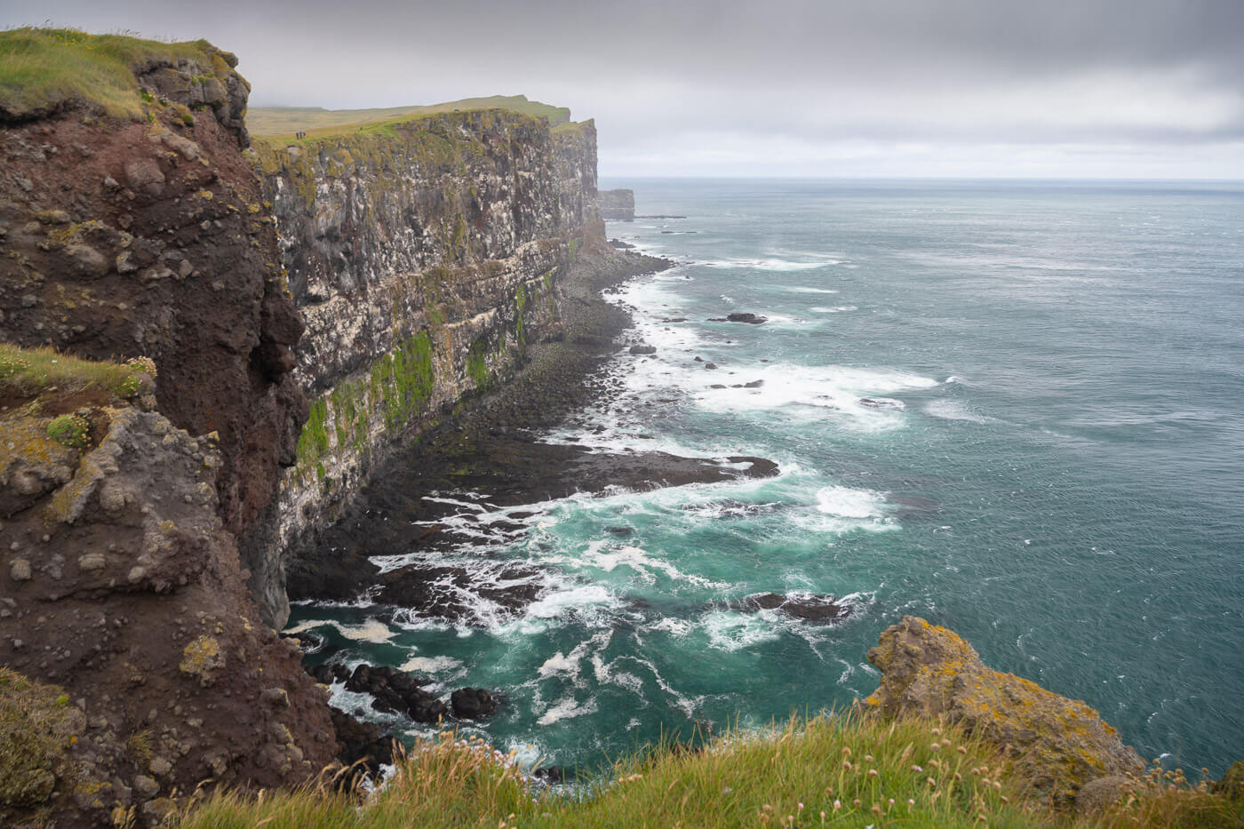 Seascape image with waves below a vertical walls above the coastline, and more cliffs in the distance.