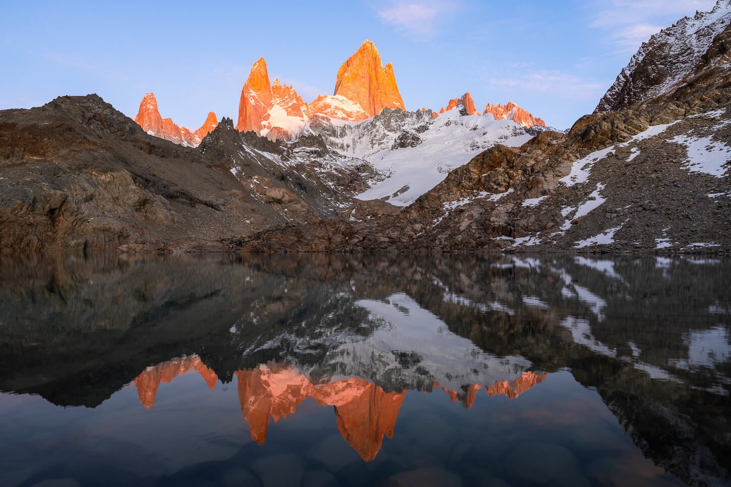 Mount Fitz Roy illuminated by the sunrise light, at the viewpoint on the shores of Laguna de Los Tres, with the mountain reflecting in the still water.