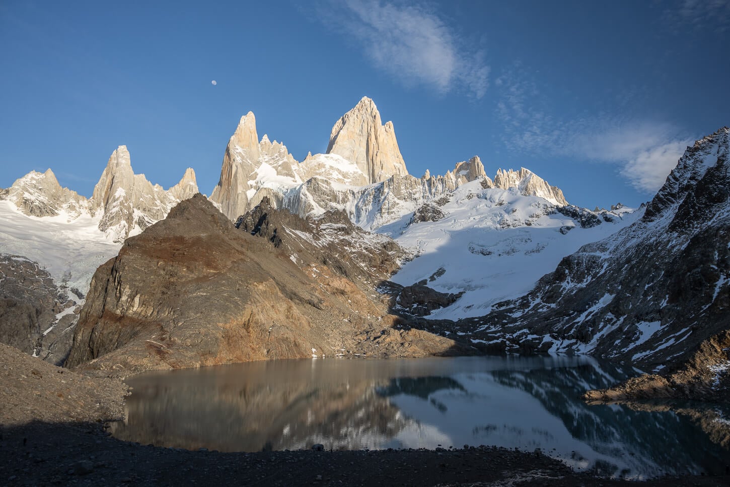 Viewpoint of Laguna the lost tres, with the Fitz Roy in the background under a blue sky, with an almost full moon in the distant background.