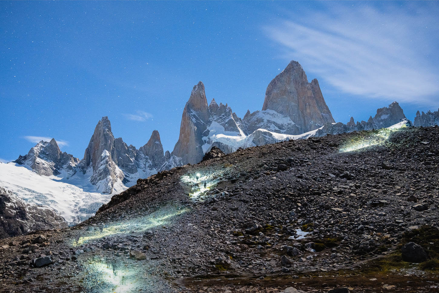 Long exposure photo of Hikers with headlamps at night, hiking up a morainic deposit to Laguna de los tres, with the Fitz Roy mountain in the background under the moonlight. 