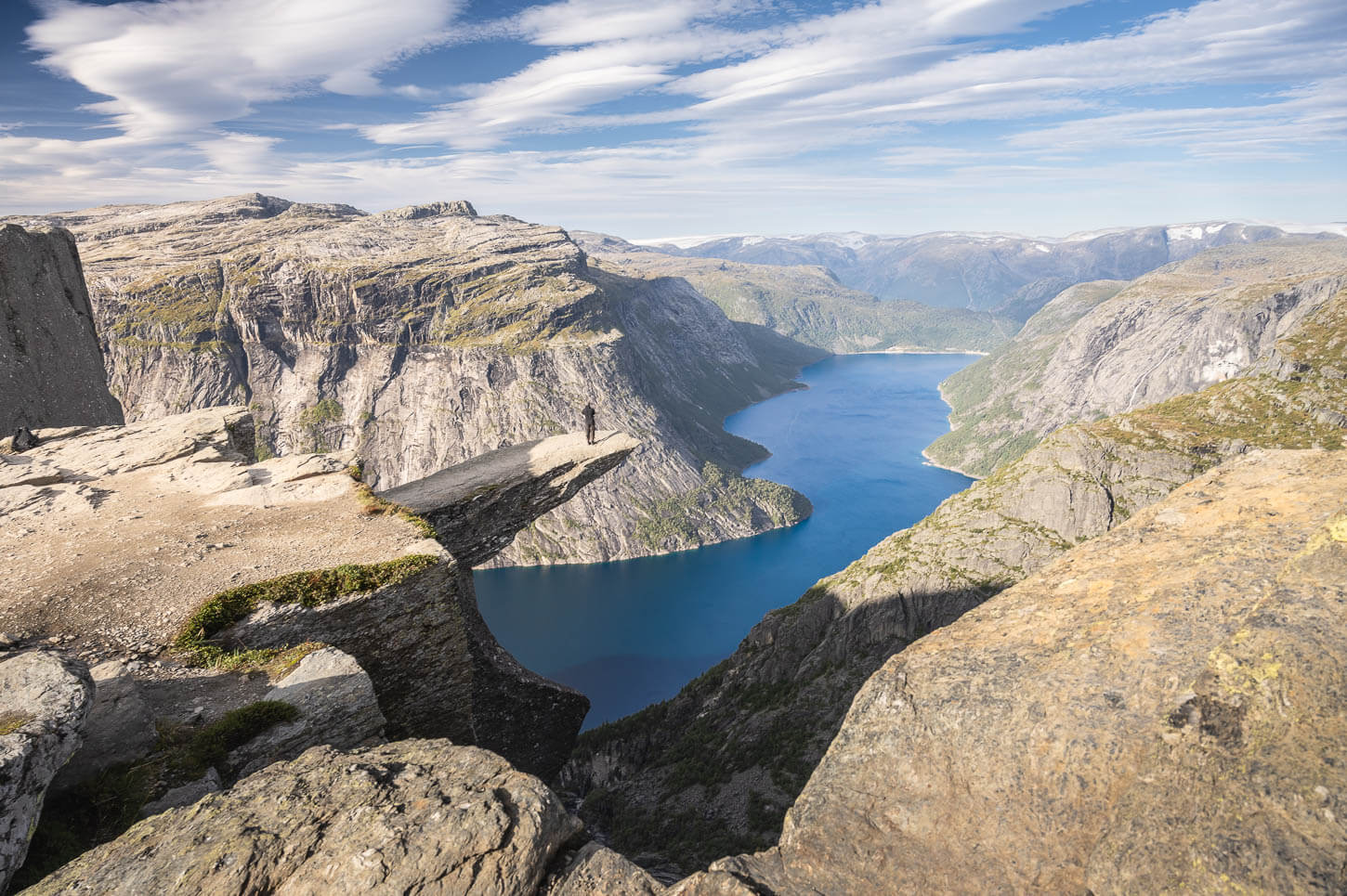 Viewpoint of Trolltunga at the end of the hike, with an hiker standing on the rock ablove the blue waters of the lake