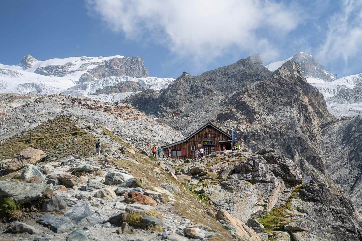 View of Rifugio Mezzalama, end point of the hike, with several glacier fronts on the background, and people around the hut.