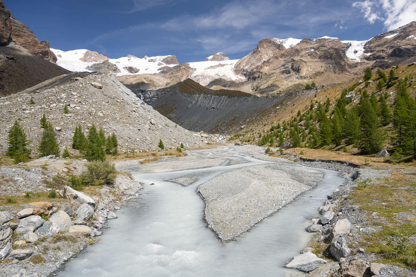 grey, silt rich waters of torrente Évançon, under a blue sky with the Monte Rosa Glacier in the background.