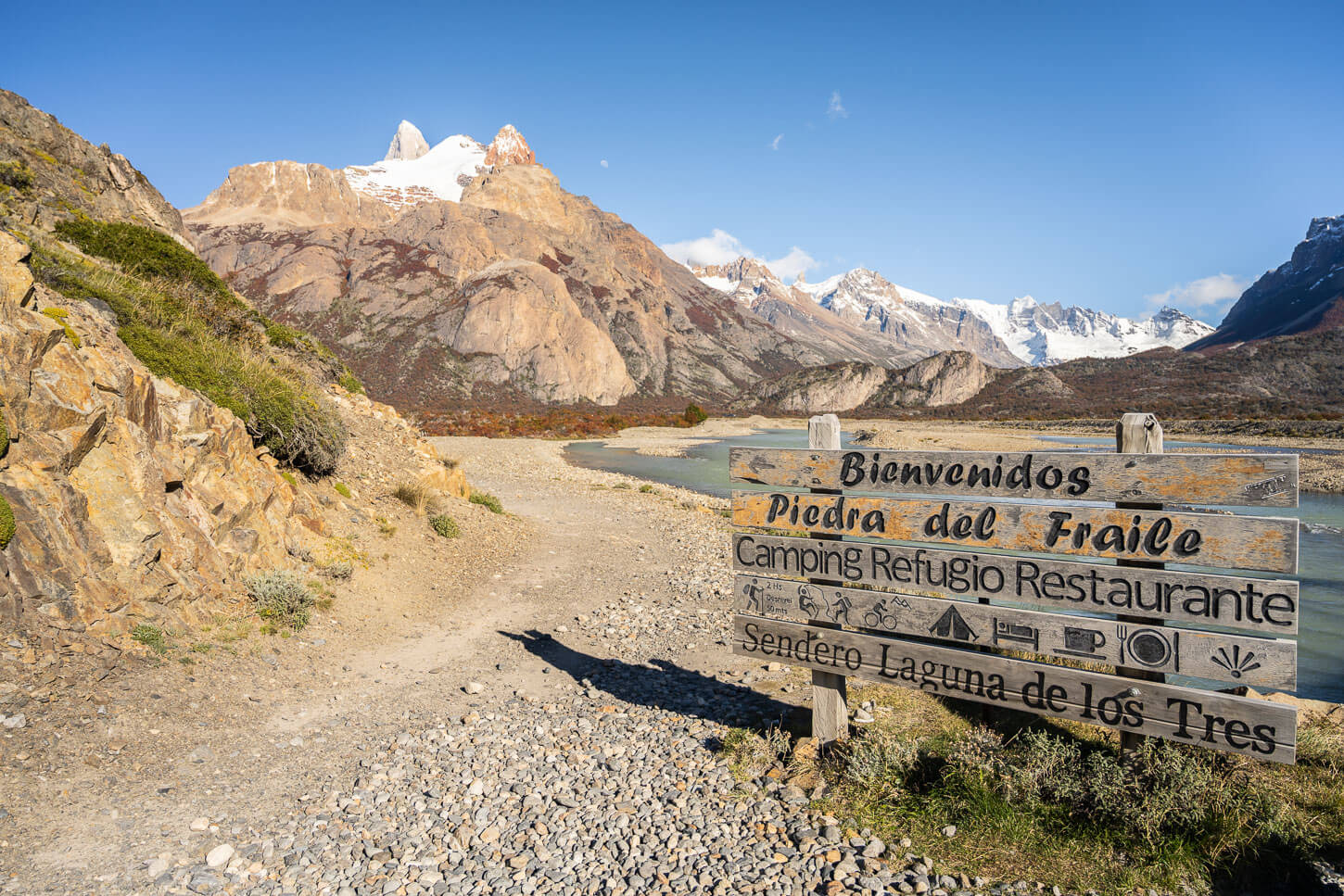 trailhead of the Mirador Piedras Blancas hike , on a sunny day, with the Rio Electrico rive in the background and the Patagonian mountains under a blue sky.