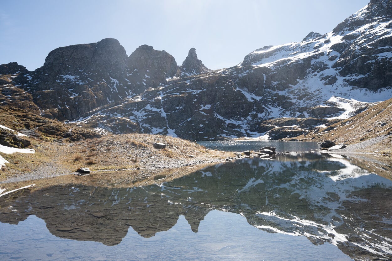 Jagged peaks reflecting in a still alpine lake, under the sun, and with snow on the mountains.