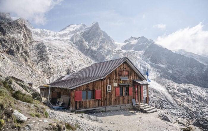 Rifugio Mezzalama on a bright sunny day, the arrival of a long hike in Val d'Ayas, with people outside the hut and glaciers perched on the mountains in the background.