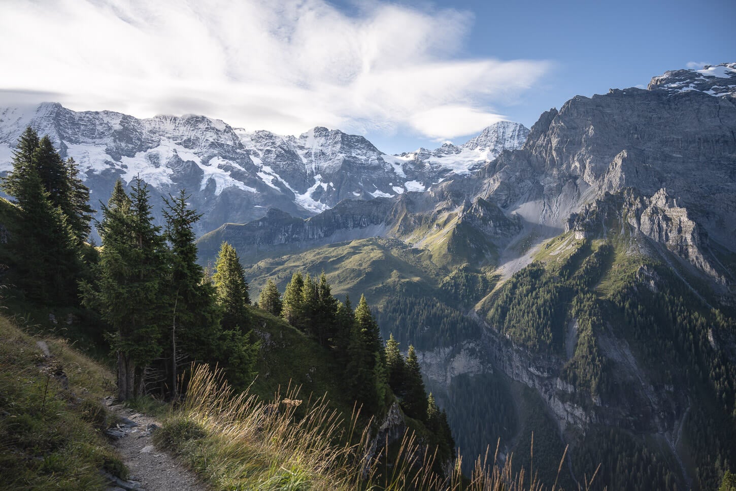 View of a hiking trail in the Bernese Oberland in the early morning, with light of the early morning coming from the east, shining on the deep, green valleys and rocky mountains.