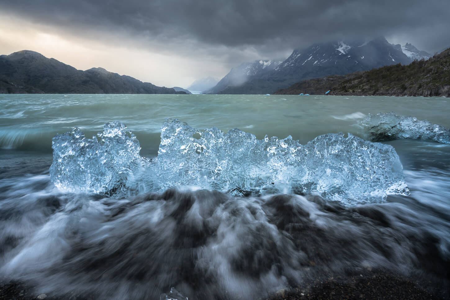 Small, shiny iceberg on the shore of Lago Grey under a dark and gloomy sky, with waves aroun d the ice.