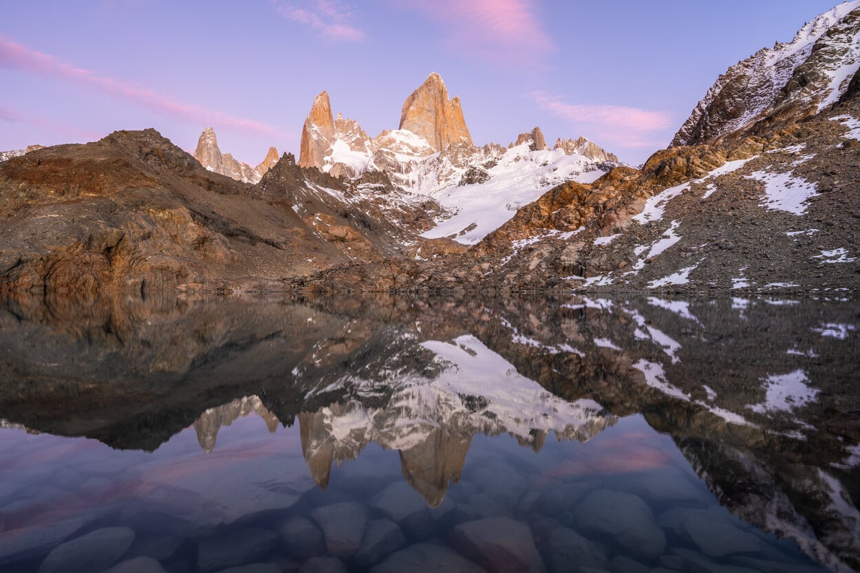 View of laguna de los Tres before sunrise, with pink hues in the sky and mountains reflecting in the water.