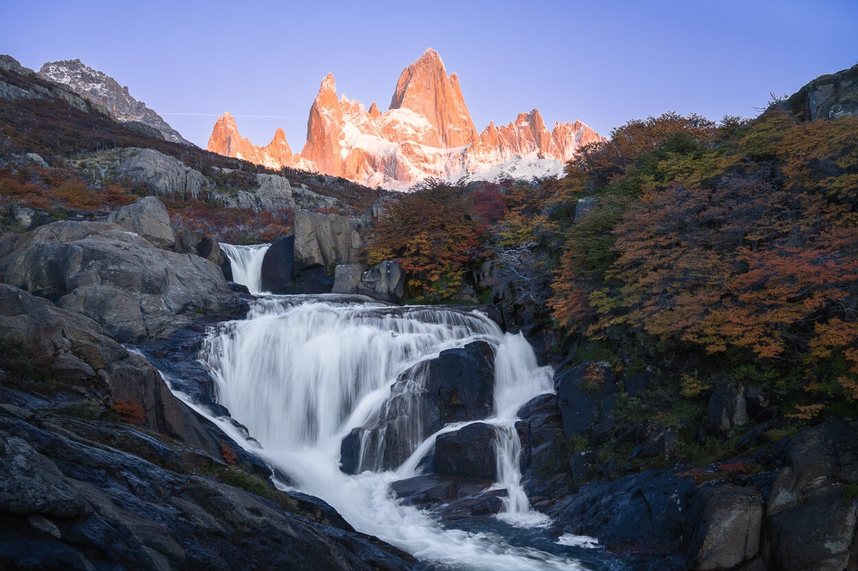 View of the hidden Waterfall in Patagonia at sunrise with the sun tinting Mount Fitz Roy in an orange-pinkish glow.