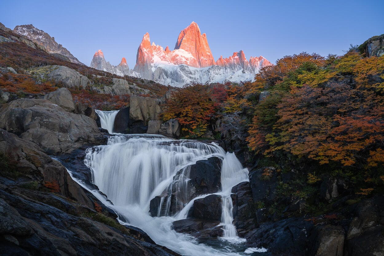View of El Chaltén's Hidden Waterfall, which makes for a spectacular sunrise hike in Patagonia, with the warm light of the rising sun hitting Mount Fitz Roy, above the hidden waterfall.
