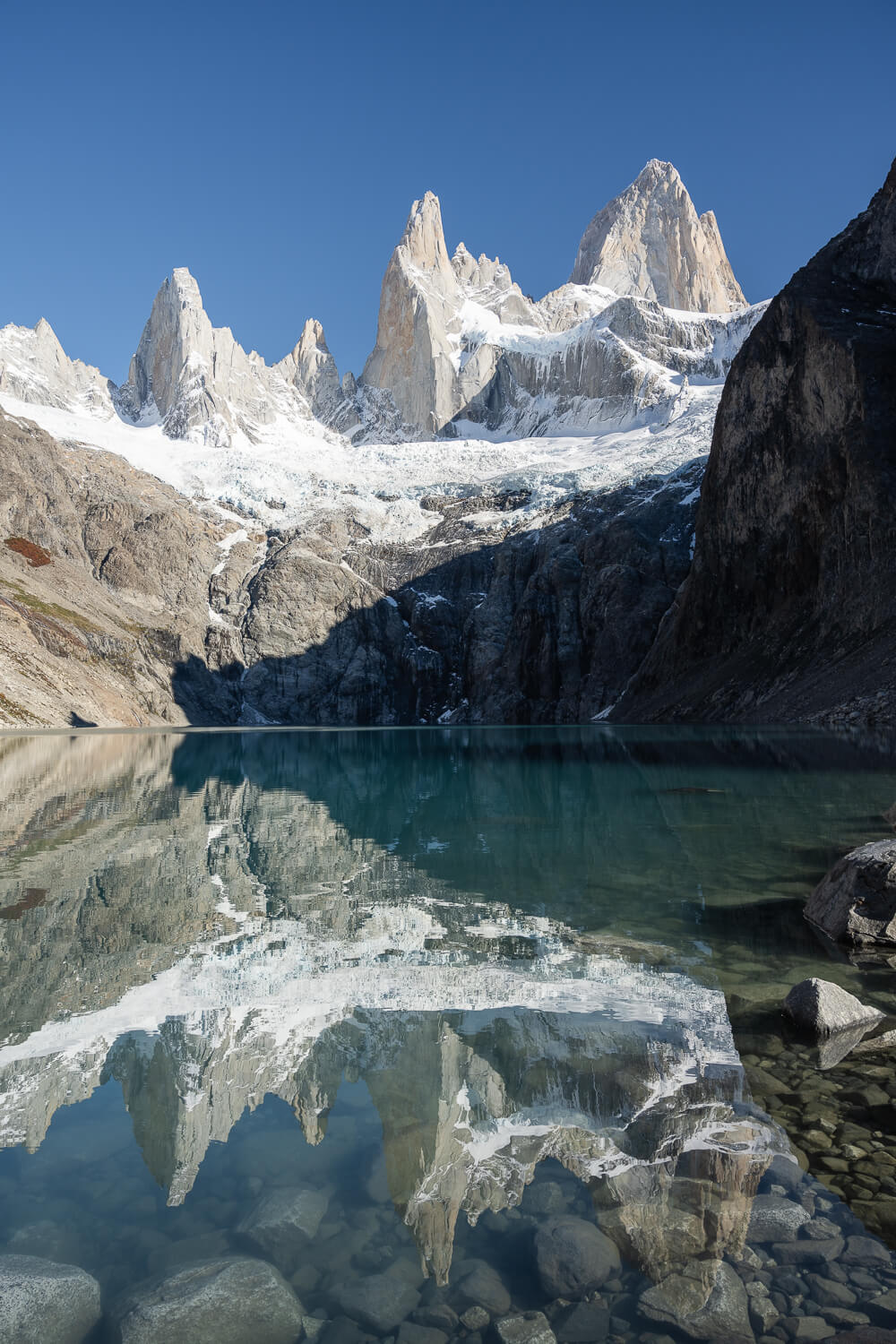 Laguna Sucia Viewpoint at the end of the hike, where mount fitz roy reflects in the calm and clear waters of the lake, on a sunny, cloudless lake.