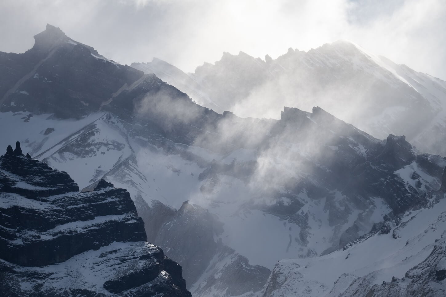 multiple layers of mountains covered in snow under some ray of light passing through the clouds.