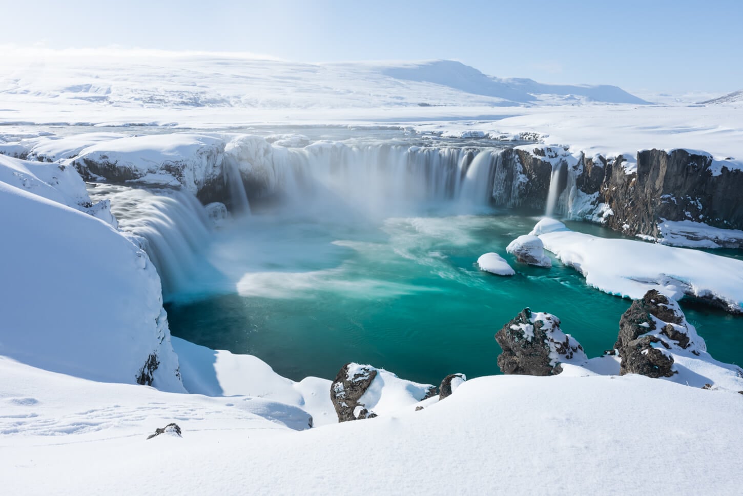 Goðafoss in Winter, partly frozen and with the surroundings covered in snow, under a sunny sky