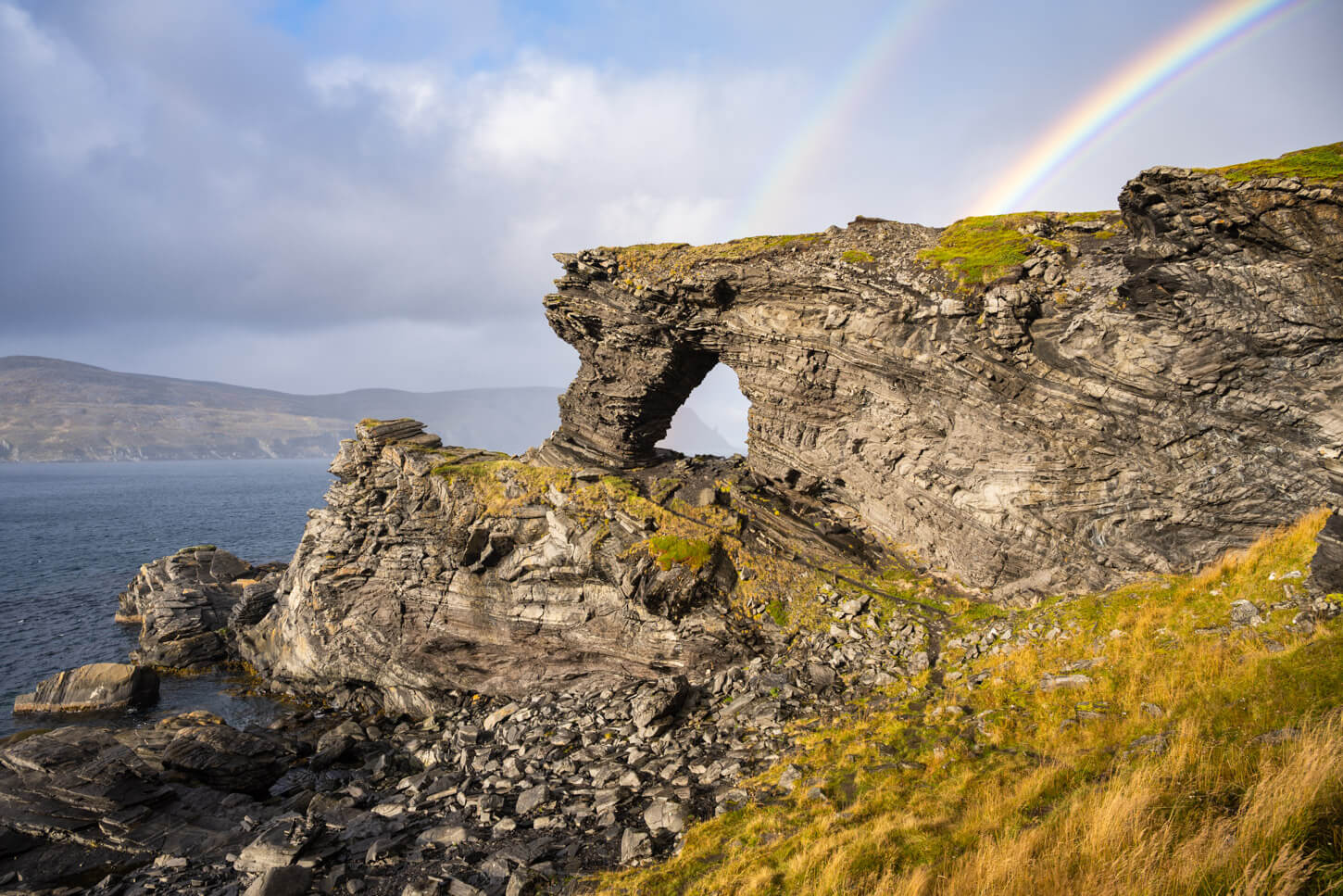 Kirkjeporten in Skarsvåg, a stone arch made of sedimentary rocks, with a raonbow above it.