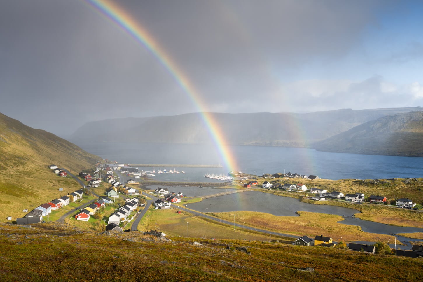 Skarsvåg under a double rainbow, with sun on one side and dark clouds on the other